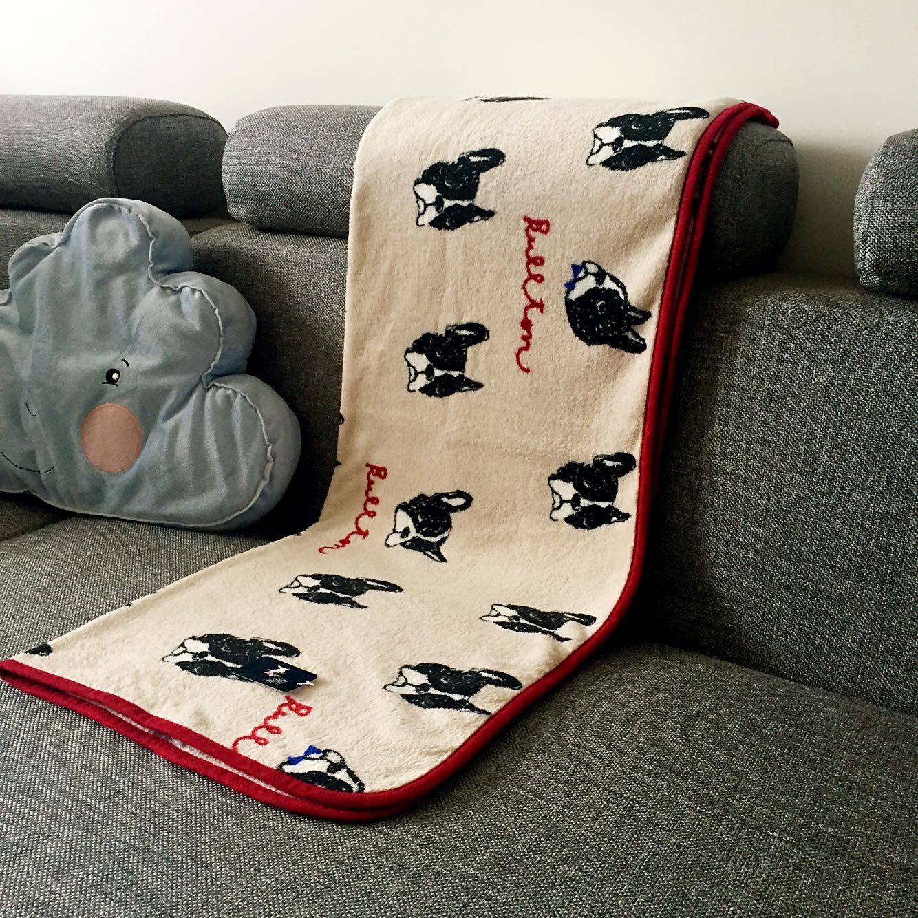 Beige pet blanket with scattered black dog faces and red border, draped on sofa